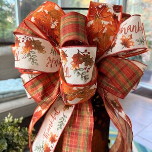 May include: A decorative bow made of wired ribbon featuring a pattern of autumn leaves, berries, and the word Thankful in cursive script on a cream background, interspersed with sections of orange, green, and brown plaid fabric ribbon.