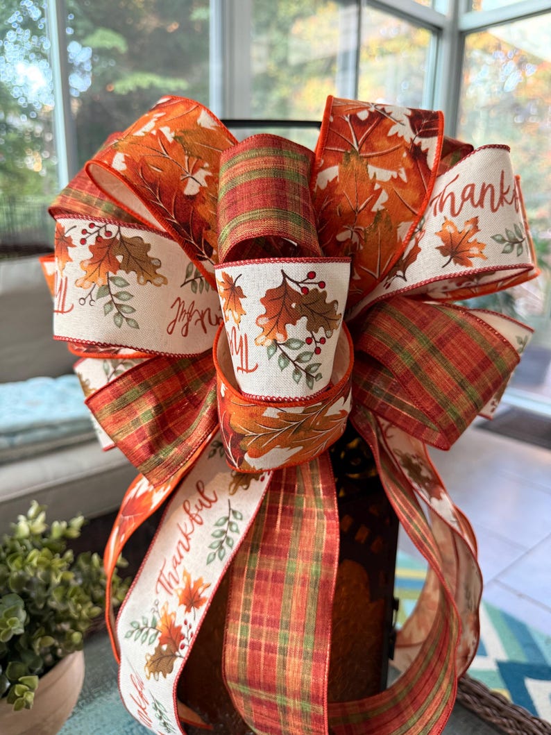 May include: A decorative bow made of wired ribbon featuring a pattern of autumn leaves, berries, and the word Thankful in cursive script on a cream background, interspersed with sections of orange, green, and brown plaid fabric ribbon.