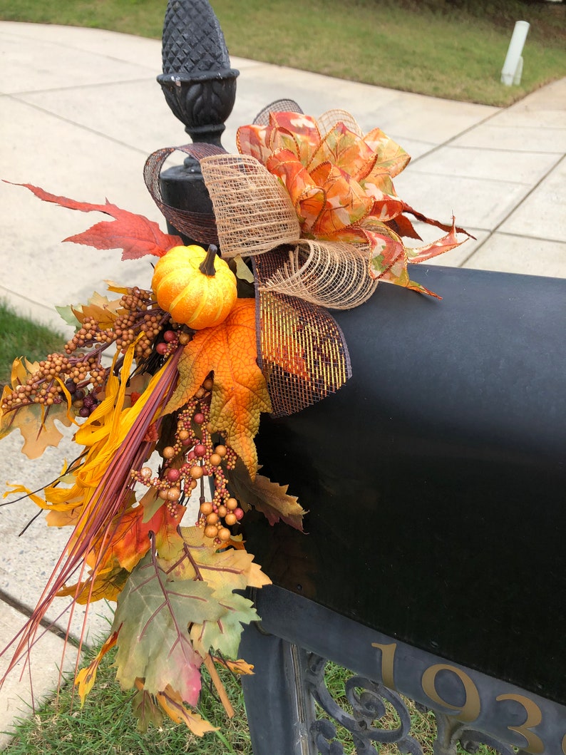 May include: A black mailbox decorated with fall-themed accents. The arrangement includes a decorative bow with orange and brown ribbons, faux pumpkins, and colorful fall leaves. The mailbox has the number 1032 in gold.