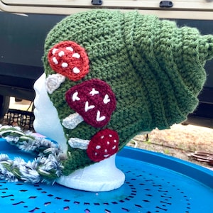 Op de afbeelding: Groene gehaakte muts met een paddenstoelontwerp. De muts heeft rode en witte paddenstoelen met witte stelen.