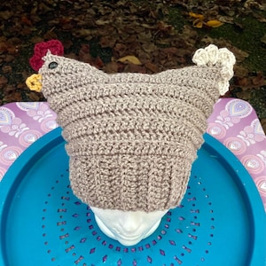 May include: A brown crocheted chicken hat with a red comb, yellow beak, and white wattle. The hat is on a styrofoam head and is sitting on a blue and purple tray.