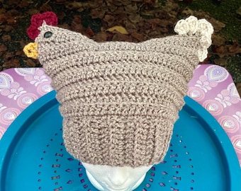 May include: A brown crocheted chicken hat with a red comb, yellow beak, and white wattle. The hat is on a styrofoam head and is sitting on a blue and purple tray.