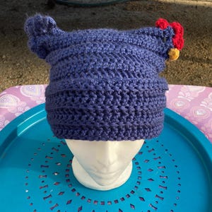 Op de afbeelding: Een blauwe gehaakte muts met een rode en gele kip-applicatie. De muts ligt op een blauw-wit gepatroneerd oppervlak.