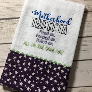 Peut inclure: Serviette de cuisine blanche avec un texte brodé qui dit "Motherhood Trifecta Peed on. Pooped on. Puked on. All on the same day." La serviette a un motif de étoiles violettes et blanches sur le bord inférieur.