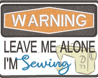 Puede incluir: Cartel bordado con el texto "WARNING" en blanco sobre un fondo naranja y gris. Debajo, se lee "LEAVE ME ALONE I'M Sewing" con un gráfico de máquina de coser. El cartel está enmarcado en negro.