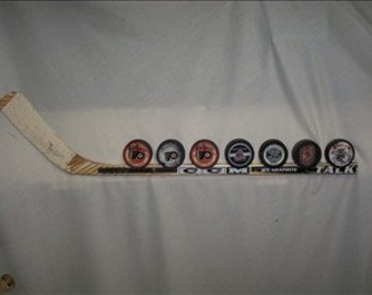 May include: A hockey stick with eight hockey pucks on it. The pucks are all different colors and have different logos on them. The stick is made of wood and has a white tape on the blade. The pucks are arranged in a row on the stick, with the logos facing the viewer.