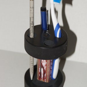 Pode incluir: Um organizador preto em forma de disco de hóquei com uma escova de dentes azul, uma caneta e um lápis com uma borracha rosa. O lápis tem o texto "Close the" impresso.