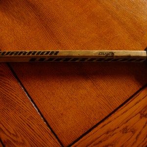 Può includere: Un bastone da hockey in legno marrone con la parola "Warrior" stampata su di esso. Il bastone è appoggiato su una superficie di legno. Un disco da hockey nero con le parole "NHL Champions" e "2019" stampate su di esso si trova a destra del bastone.