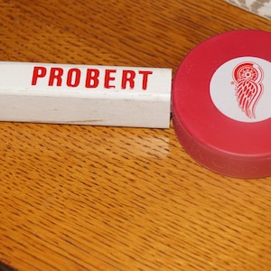 May include: A white hockey stick with the name "PROBERT" written in red letters. The stick is attached to a red hockey puck with a white Detroit Red Wings logo.