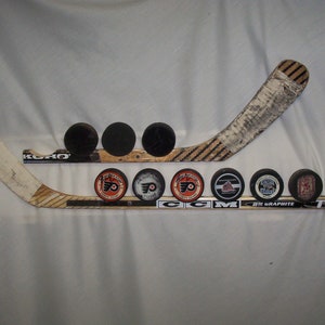 May include: A hockey stick with a white blade and a black and brown shaft. The stick is mounted on a wall with a black and brown hockey stick. There are nine hockey pucks on the wall, some with logos and some plain black.