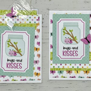May include: Two handmade greeting cards with a green, white, and pink color scheme. The cards feature a jar with bugs and the text "bugs and kisses".