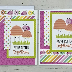 May include: Two pink and orange mushroom illustrations with smiling faces and ladybug accents on a pink and green card with the text "We're better together".