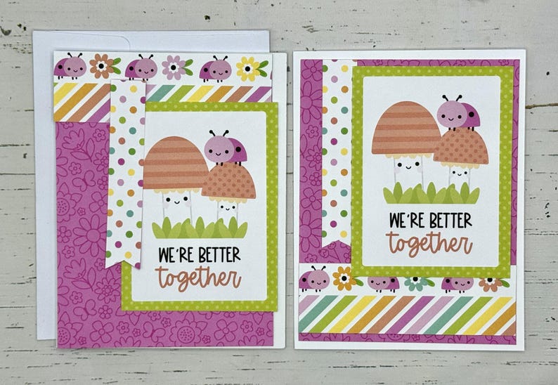May include: Two pink and orange mushroom illustrations with smiling faces and ladybug accents on a pink and green card with the text "We're better together".
