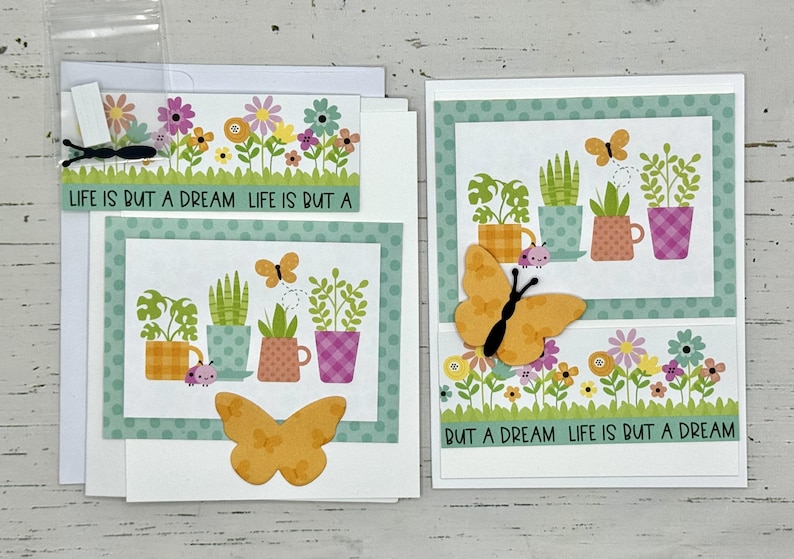 May include: Two handmade greeting cards with a green and white polka dot border. The cards feature a floral design with potted plants and butterflies. The text on the cards reads "Life is but a dream".