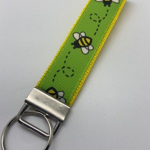 May include: Green and yellow key chain with a bee design. The key chain has a silver key ring.