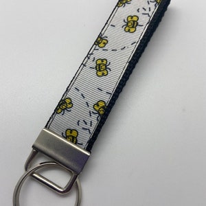 May include: A white and black key chain with a silver key ring. The white strap has a repeating pattern of yellow bees.