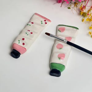 Può includere: Due portatubi in ceramica a forma di tubetto di vernice, uno con ciliegie e l'altro con fragole. I tubi sono bianchi con accenti rosa e verdi e tappi neri. Un pennello poggia sul tubetto di fragole.
