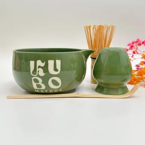 以下が含まれることがあります： KUBO MATCHAの白い文字が書かれたセージグリーンの陶器製抹茶碗、竹製の茶筅、緑色の陶器製茶筅立て、長い竹製の茶杓。抹茶碗の右側には注ぎ口があり、無地の表面に配置されています。