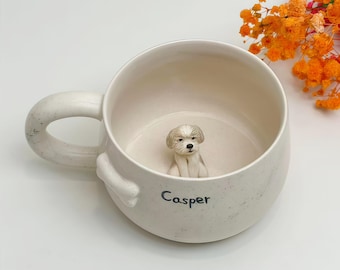 May include: A cream-colored ceramic mug with a speckled finish, featuring a small, sculpted dog figure inside at the base. The exterior displays the name Casper in black lettering and a bone-shaped detail near the handle attachment point.