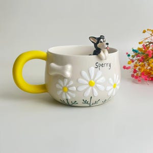Puede incluir: Una taza de cer&aacute;mica beige con un asa amarilla brillante, decorada con margaritas blancas en relieve, un peque&ntilde;o hueso blanco y el nombre Sperry escrito en negro. Una peque&ntilde;a figura de perro blanco y negro est&aacute; posada en el borde.