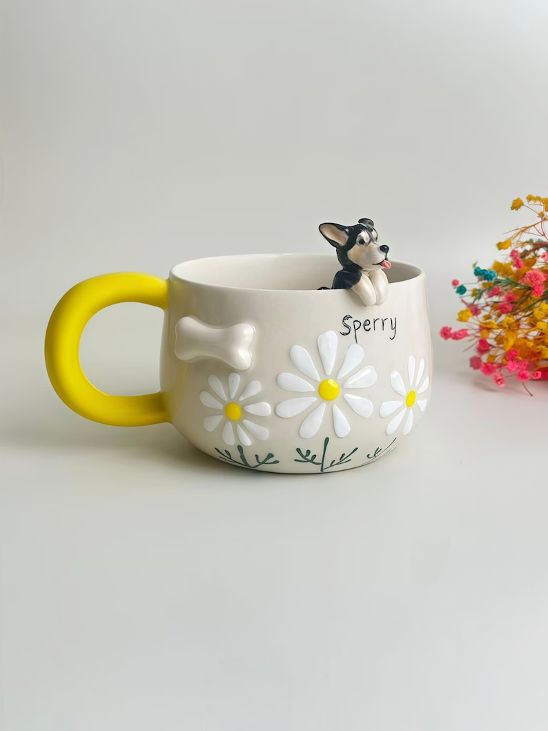 Puede incluir: Una taza de cer&aacute;mica beige con un asa amarilla brillante, decorada con margaritas blancas en relieve, un peque&ntilde;o hueso blanco y el nombre Sperry escrito en negro. Una peque&ntilde;a figura de perro blanco y negro est&aacute; posada en el borde.