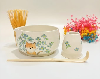 May include: A ceramic matcha bowl featuring a painted orange cat and blue flowers, a matching whisk holder, a bamboo whisk, and a bamboo tea scoop. The bowl has a small pouring spout on the side, and the set is decorated with a consistent floral motif.