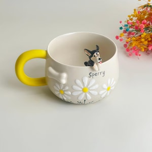 Puede incluir: Una taza de cer&aacute;mica color crema con un asa amarilla brillante, que presenta una figura tridimensional de un perro en blanco y negro asom&aacute;ndose por el borde. La taza est&aacute; decorada con margaritas blancas, un hueso blanco y el nombre Sperry en negro.