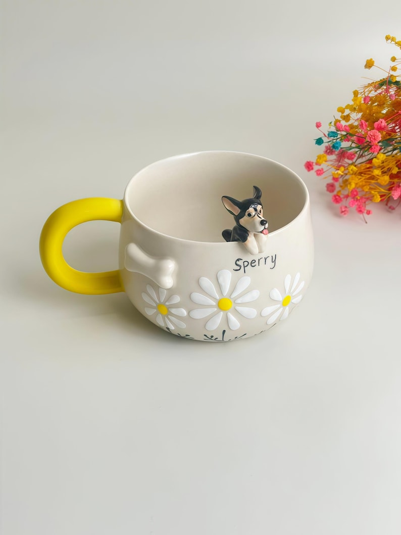 Puede incluir: Una taza de cer&aacute;mica color crema con un asa amarilla brillante, que presenta una figura tridimensional de un perro en blanco y negro asom&aacute;ndose por el borde. La taza est&aacute; decorada con margaritas blancas, un hueso blanco y el nombre Sperry en negro.