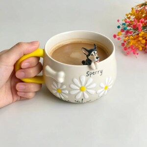 Puede incluir: Una taza de cer&aacute;mica color crema con asa amarilla brillante, que presenta una figura de perro en 3D en blanco y negro sobre el borde, patrones de margaritas blancas, un detalle en relieve con forma de hueso y el nombre Sperry en letras negras.