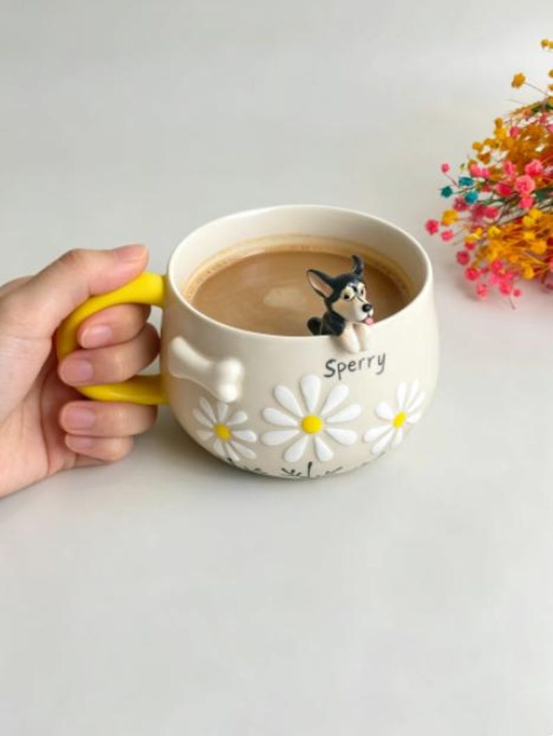 Puede incluir: Una taza de cer&aacute;mica color crema con asa amarilla brillante, que presenta una figura de perro en 3D en blanco y negro sobre el borde, patrones de margaritas blancas, un detalle en relieve con forma de hueso y el nombre Sperry en letras negras.