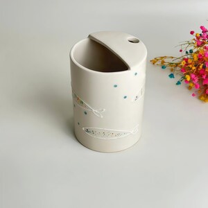 May include: A cylindrical off-white ceramic utensil holder featuring a slanted top opening and a small circular hole. The exterior is decorated with hand-painted white fish outlines filled with tiny multicolored dots and scattered blue dots.