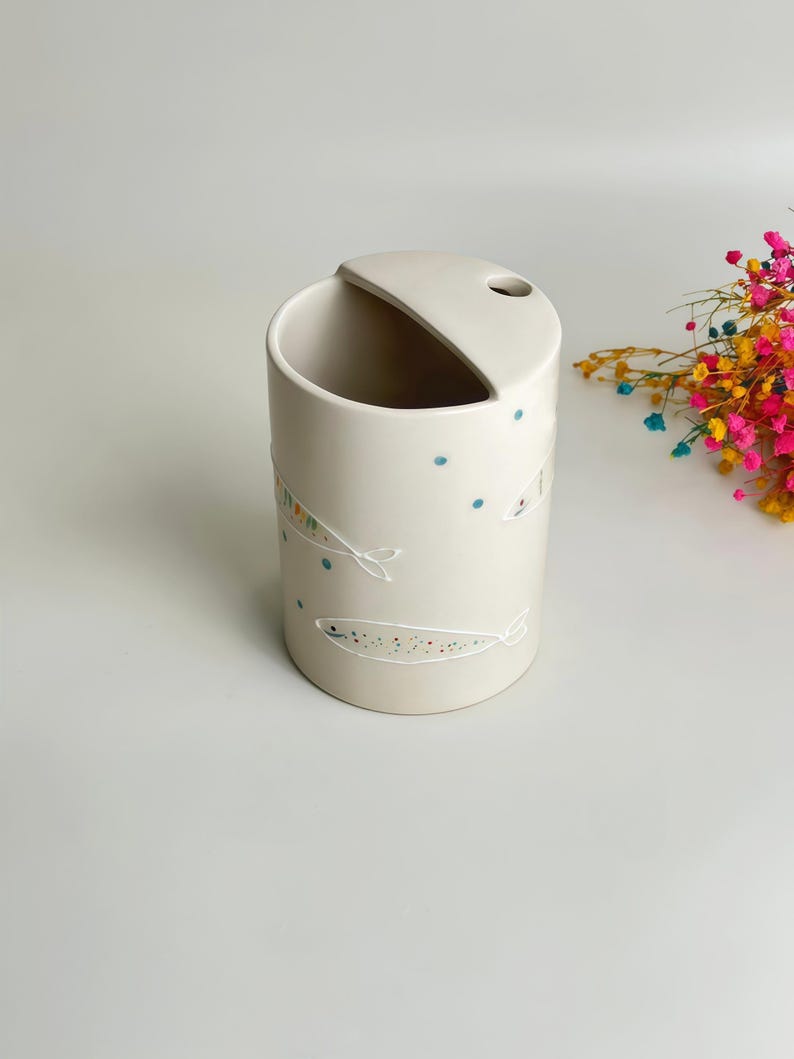 May include: A cylindrical off-white ceramic utensil holder featuring a slanted top opening and a small circular hole. The exterior is decorated with hand-painted white fish outlines filled with tiny multicolored dots and scattered blue dots.