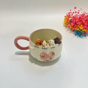 以下が含まれることがあります： ピンクの持ち手と縁取りが施されたクリーム色の陶器製マグカップ。内側の縁には4体の小さなモルモットの彫刻が飾られています。彫刻の下には「BERTI」「MIKE」「LAIA」という名前が印字されており、白い花が描かれたピンクのハートの装飾が施されています。