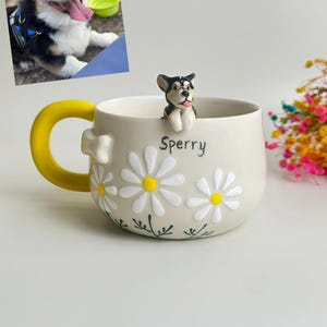 Puede incluir: Una taza de cer&aacute;mica con base color crema, asa amarilla brillante y aplicaciones de margaritas blancas con centros amarillos. Una peque&ntilde;a figura esculpida de un perro negro, blanco y marr&oacute;n se asienta en el borde. El nombre Sperry est&aacute; impreso.