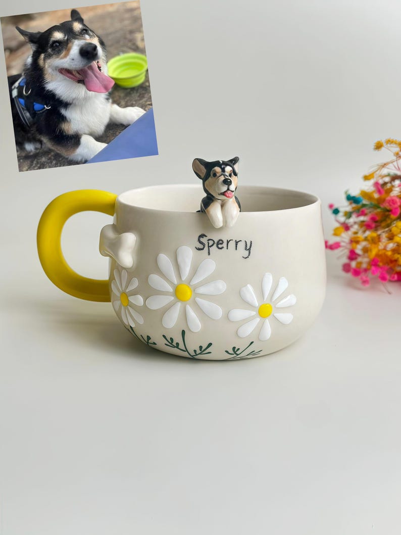 Puede incluir: Una taza de cer&aacute;mica con base color crema, asa amarilla brillante y aplicaciones de margaritas blancas con centros amarillos. Una peque&ntilde;a figura esculpida de un perro negro, blanco y marr&oacute;n se asienta en el borde. El nombre Sperry est&aacute; impreso.