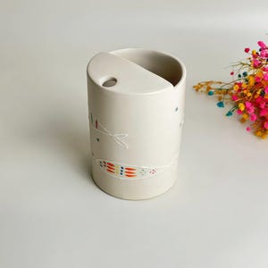 May include: A cylindrical off-white ceramic holder featuring a semi-circular top opening with a small circular hole. The exterior is decorated with delicate white line drawings of fish, accented with small, colorful dots in red, orange, and blue.