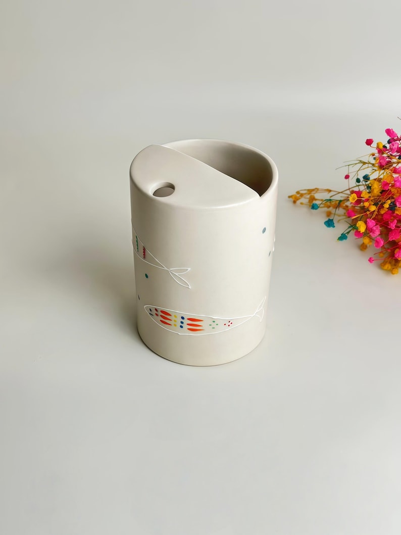 May include: A cylindrical off-white ceramic holder featuring a semi-circular top opening with a small circular hole. The exterior is decorated with delicate white line drawings of fish, accented with small, colorful dots in red, orange, and blue.