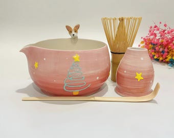 May include: A pink ceramic matcha bowl with a small dog figurine on the rim, featuring a painted Christmas tree and stars. Includes a bamboo whisk, a bamboo scoop, and a pink ceramic whisk holder, all decorated with white dots and yellow stars.