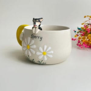 Puede incluir: Una taza de cer&aacute;mica beige con una figura de perro esculpida en blanco y negro posada en el borde. La taza est&aacute; decorada con margaritas blancas de centro amarillo y el nombre Sperry escrito en negro. Tiene un asa de color amarillo brillante.