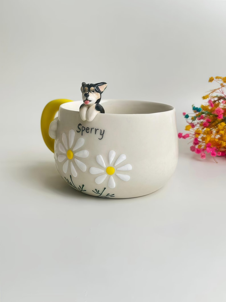 Puede incluir: Una taza de cer&aacute;mica beige con una figura de perro esculpida en blanco y negro posada en el borde. La taza est&aacute; decorada con margaritas blancas de centro amarillo y el nombre Sperry escrito en negro. Tiene un asa de color amarillo brillante.