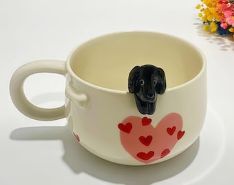 Puede incluir: Taza de cerámica color crema con una figura de perro negro en el borde y un diseño de corazón rosa con corazones rojos. La taza tiene un asa curva y es adecuada para café, té u otras bebidas.