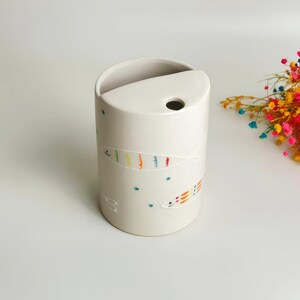 May include: A cylindrical off-white ceramic vessel featuring a semi-circular top opening and a small circular hole. The exterior is decorated with hand-painted outlines of fish in vibrant rainbow colors, interspersed with small teal dots.