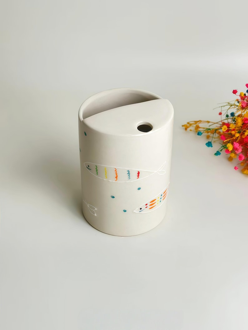 May include: A cylindrical off-white ceramic vessel featuring a semi-circular top opening and a small circular hole. The exterior is decorated with hand-painted outlines of fish in vibrant rainbow colors, interspersed with small teal dots.