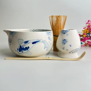 Puede incluir: Un cuenco de cerámica para matcha con un patrón de carpa koi y olas azules, un batidor de bambú, un soporte de cerámica para el batidor con motivos florales azules y una cuchara de bambú larga. El cuenco tiene un pico vertedor y acabado esmaltado.