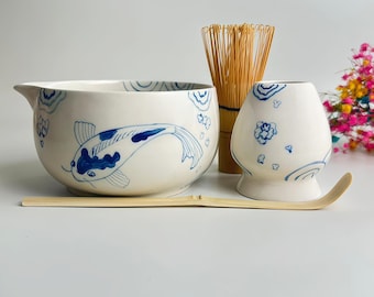 May include: A ceramic matcha bowl with a blue koi fish and wave pattern, a bamboo whisk, a ceramic whisk holder with blue floral motifs, and a long bamboo scoop. The bowl features a pouring spout and a smooth, off-white glazed finish.