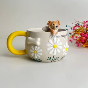 Puede incluir: Una taza de cer&aacute;mica beige con un asa amarilla brillante, que presenta una figura esculpida de un perro marr&oacute;n y blanco en el borde. La taza est&aacute; decorada con margaritas blancas en relieve, tallos pintados en verde y un hueso blanco. El texto Chicken Nugget est&aacute; impreso.