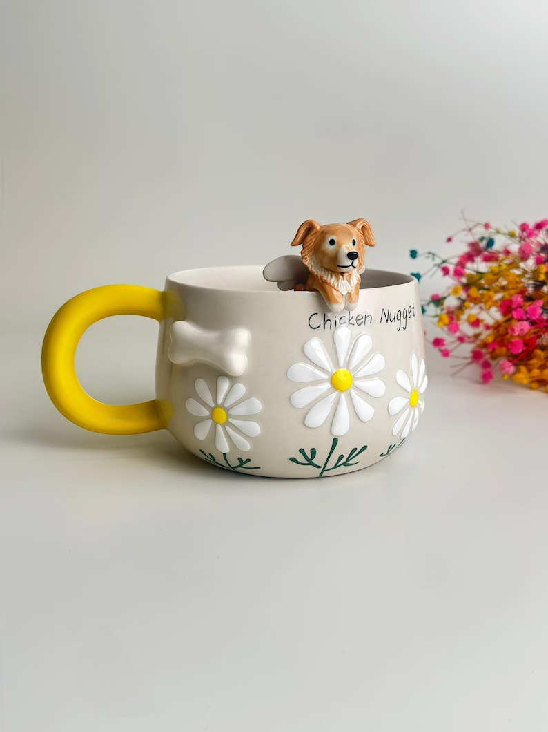 Puede incluir: Una taza de cer&aacute;mica beige con un asa amarilla brillante, que presenta una figura esculpida de un perro marr&oacute;n y blanco en el borde. La taza est&aacute; decorada con margaritas blancas en relieve, tallos pintados en verde y un hueso blanco. El texto Chicken Nugget est&aacute; impreso.