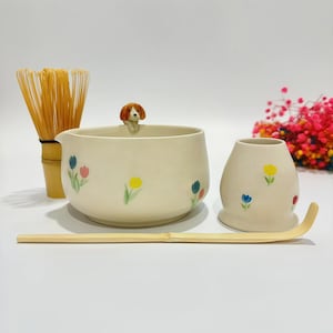 May include: A cream-colored ceramic matcha tea set with floral accents, including a bowl with a dog figurine, a whisk, a scoop, and a small cup. The set features hand-painted flowers in various colors.