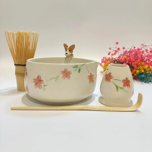 Könnte beinhalten: Ein Matcha-Teeset mit einer Keramikschale, einem Schneebesen, einem Löffel und einem kleinen Behälter. Schale und Behälter sind mit rosa floralen Designs verziert. Eine kleine Hundefigur sitzt am Rand der Schale. Das Set enthält einen Bambusbesen und -löffel.