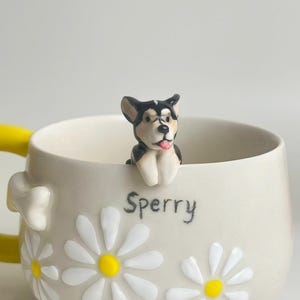 Puede incluir: Una taza de cer&aacute;mica color crema con una figura tridimensional de un perro en blanco y negro asom&aacute;ndose por el borde. La taza est&aacute; decorada con p&eacute;talos de margarita blancos en relieve con centros amarillos y el nombre Sperry en negro.